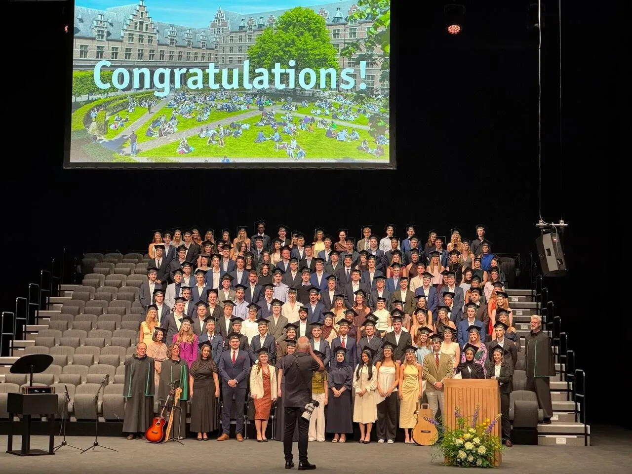 Graduates in caps and gowns on stage under a screen reading "Congratulations!"